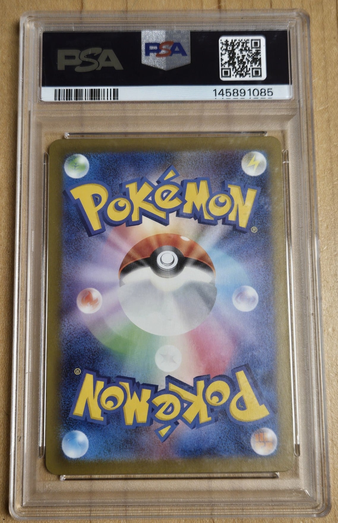 A PSA graded Pokémon card featuring a colorful Pokémon logo and a Pokémon Ball design on the back, sealed inside a clear PSA slab on a light wood surface.
