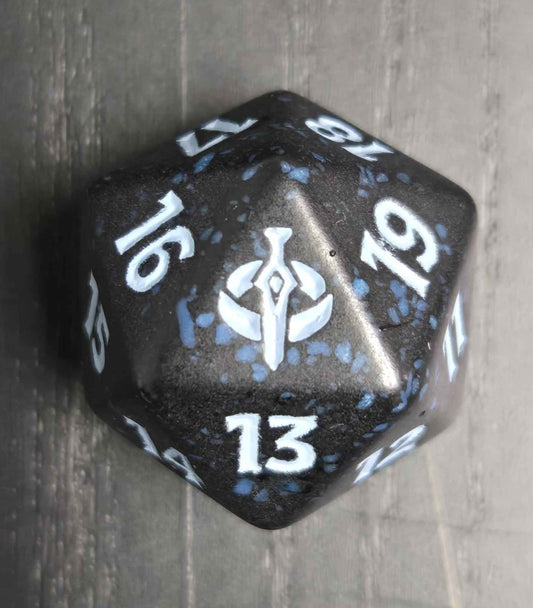 A black polyhedral die with blue marbled speckles and white printed numbers and a central symbol, photographed on a gray wood-textured surface.