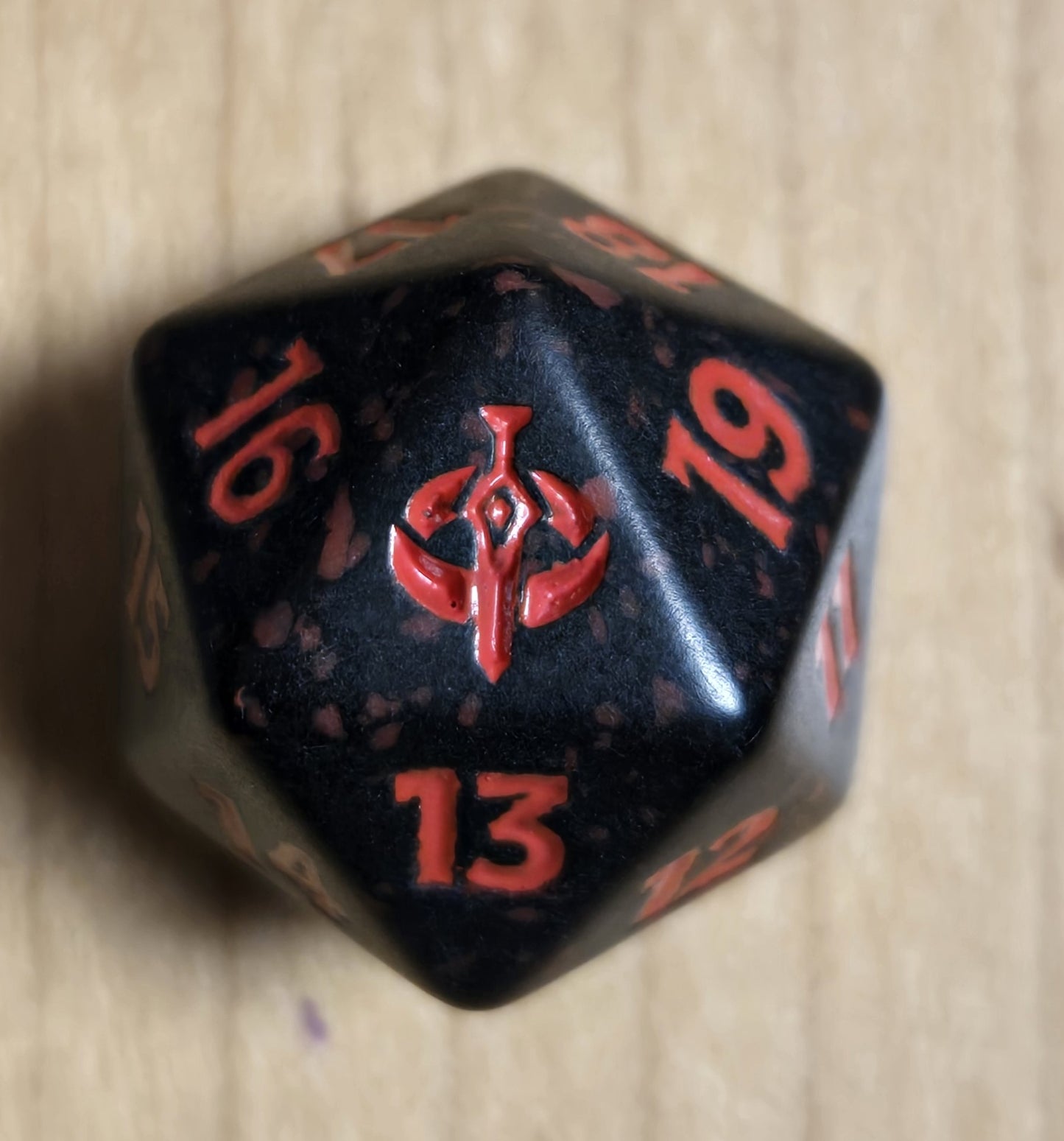 A black polyhedral gaming die with red numbering and a red central symbol, photographed on a light wood surface.